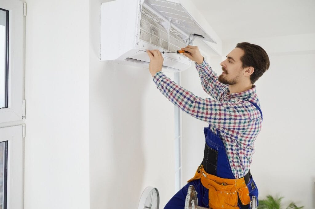 Smiling young male electrician repair conditioner in client home. Man mechanic or repairman fix filters in air condition device at customer house. Maintenance and household help concept.